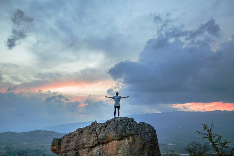 Man worshiping God with raised arms while staring out at the heavens on top of a rock